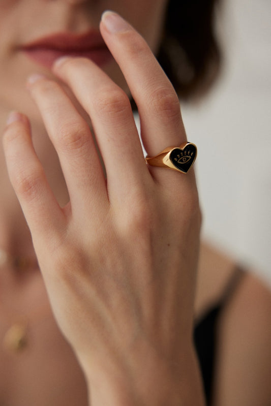 A close-up image of a person's hand wearing the Vintage Gold Adjustable Evil Eye Ring in 925 Silver with Enamel Detail by Siren Mode on their ring finger. The ring has a black face with an inscription. The person's mouth is partially visible, and they are lightly touching their lips with the ring-wearing hand.