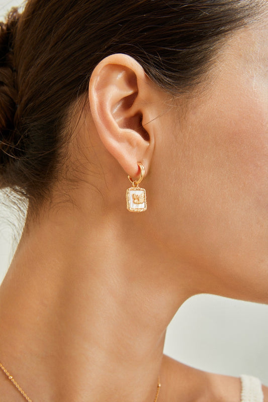 Close-up of a woman's left ear showcasing the Siren Mode Vintage Gold Butterfly Pendant Earring with a gemstone at its center. The woman, wearing a white top, has short brown hair tied back. These stunning gold-plated earrings add an elegant touch to her look. The background is blurred and neutral in color.