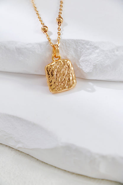 A close-up of the exquisite Vintage Gold Butterfly Pendant Necklace by Siren Mode showcases its textured, rectangular charm. The high-quality gold-plated chain, adorned with small gold beads spaced along it, enhances its elegance. The necklace is beautifully displayed on a white, textured surface.