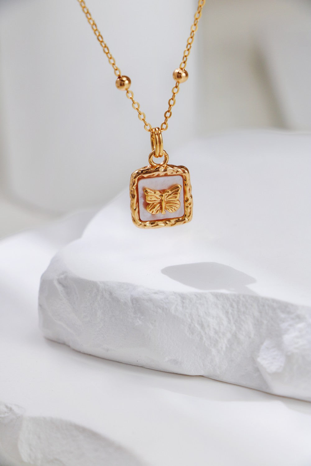 A delicate, high-quality Vintage Gold Butterfly Pendant Necklace by Siren Mode is displayed on a white textured surface. The rectangular pendant features a butterfly design, while the gold chain has small beads spaced evenly along it, enhancing the elegant and intricate look of this exquisite necklace.