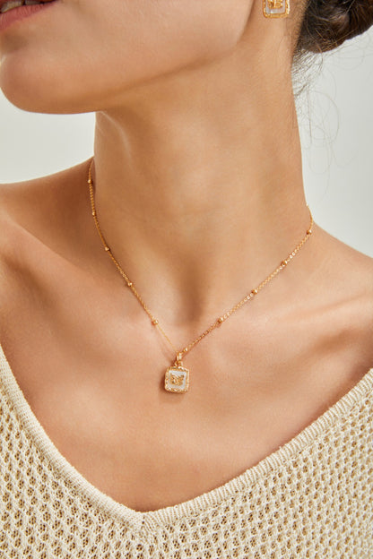 Close-up of a person wearing an exquisite Vintage Gold Butterfly Pendant Necklace by Siren Mode, featuring a square charm encrusted with small stones. The person is also wearing a crocheted, off-white top, and the background is softly blurred, drawing focus to the high-quality gold-plated jewelry.