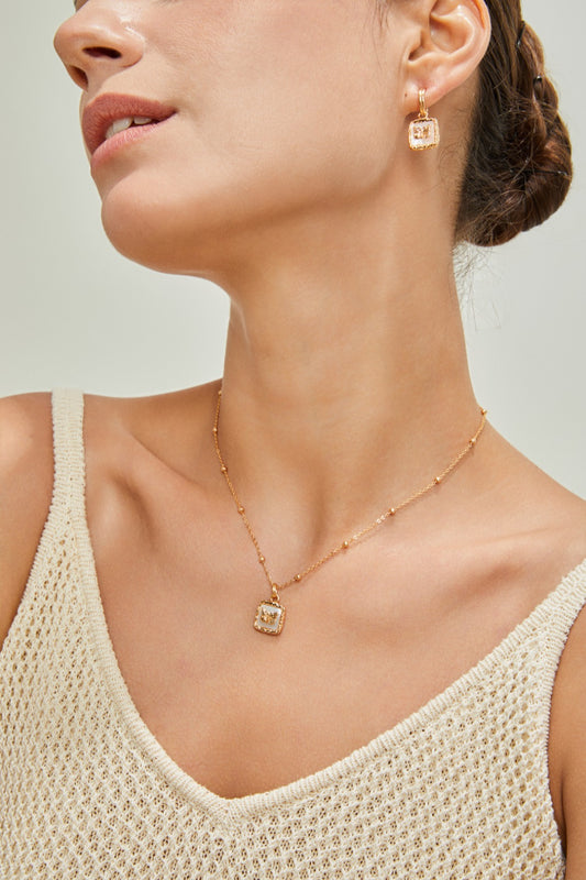 A woman with her hair pulled back is wearing the Siren Mode Vintage Gold Butterfly Pendant Necklace and matching earrings, each featuring a square-shaped pendant with a detailed design. She is dressed in a light, knitted top and is gazing towards the upper left side of the image.