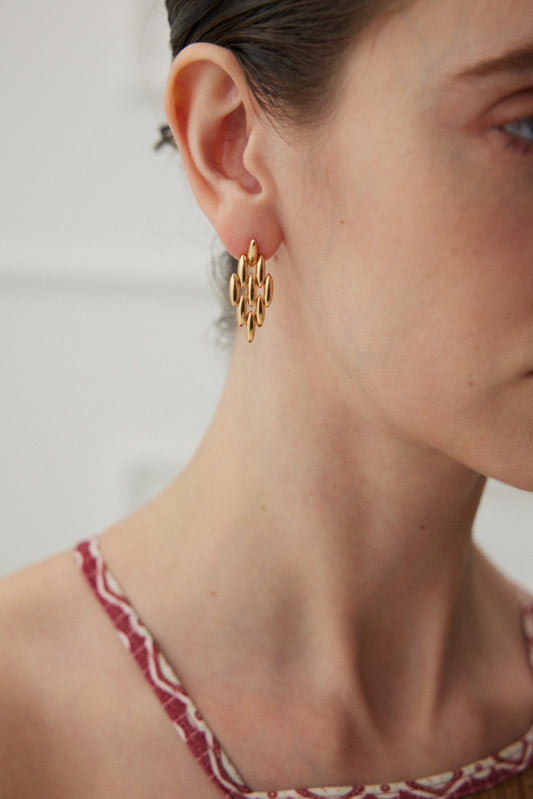 Close-up of a person wearing Siren Mode's Vintage Gold Chain Link Drop Earrings in 925 Silver. They are also wearing a patterned, red and white sleeveless top. The person's hair is pulled back, and the background is softly blurred.