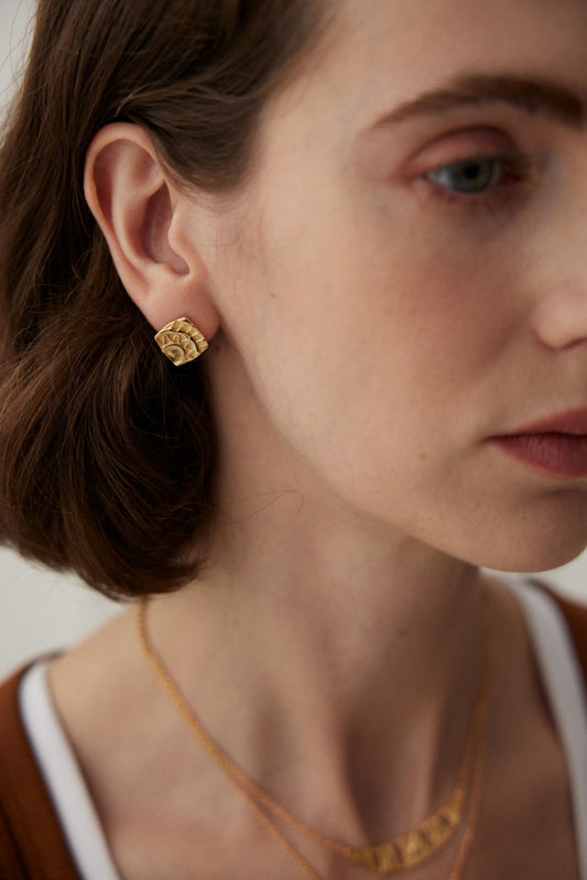 Close-up of a woman wearing a pair of Siren Mode's Vintage Gold Geometric Stud Earrings shaped like folded pieces of paper. She also has a delicate gold necklace, and her brown hair is tucked behind her ear. The background is softly lit, emphasizing the jewelry's geometric pattern.