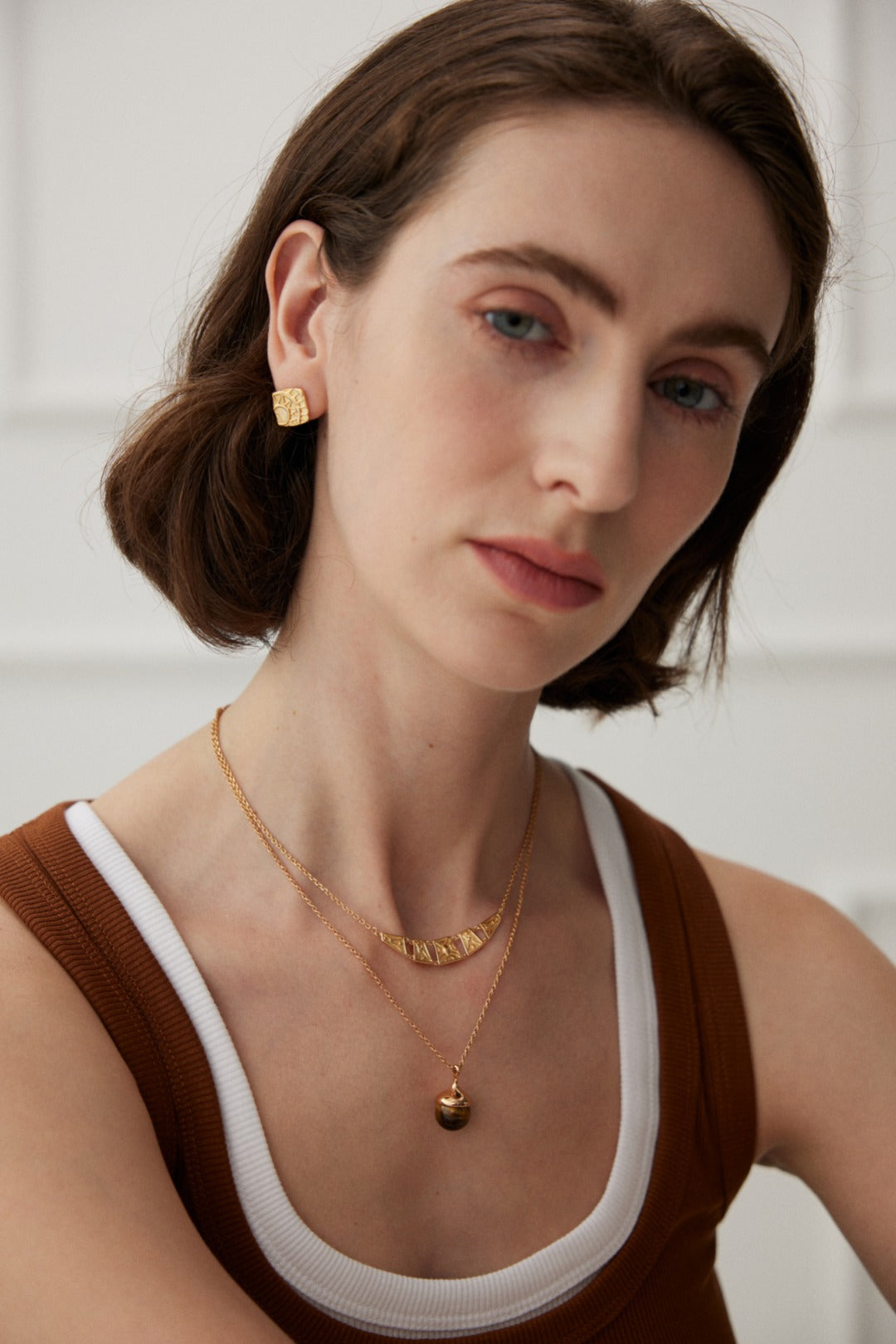 A woman with short brown hair and light skin is wearing a sleeveless top featuring a brown and white geometric pattern. She accessorizes with Siren Mode's Vintage Gold Geometric Stud Earrings, accompanied by two gold necklaces – one adorned with a small pendant and the other showcasing lettered charms. She gazes softly at the camera against a blurred, white background.