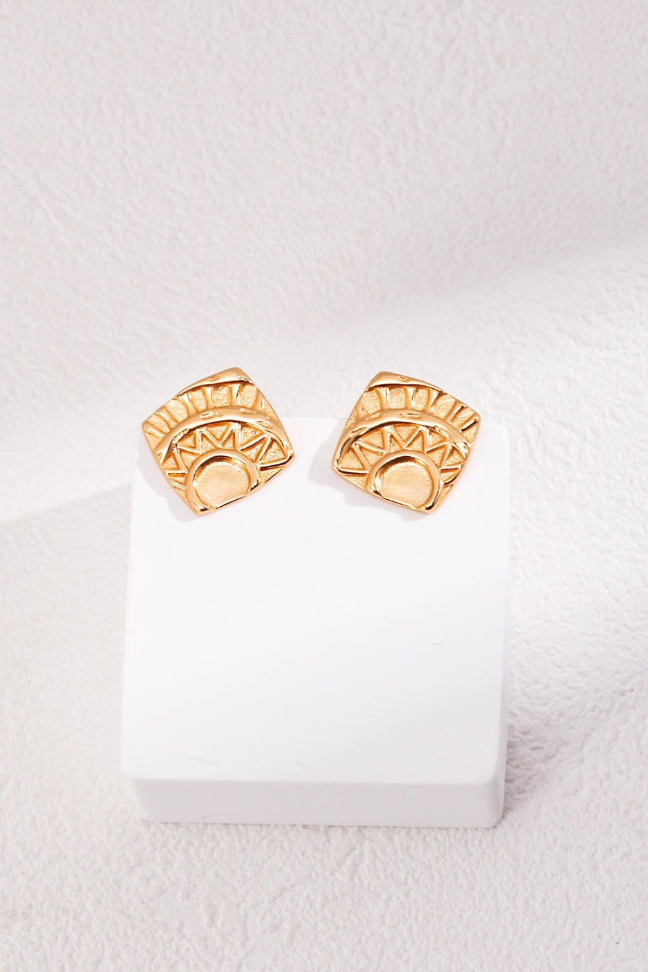 The Vintage Gold Geometric Stud Earrings by Siren Mode feature engraved sun motifs on square-shaped studs crafted from 925 silver. Displayed elegantly on a white, textured surface and perched on a small white pedestal, the intricate design showcases geometric patterns that beautifully complement the sun imagery.