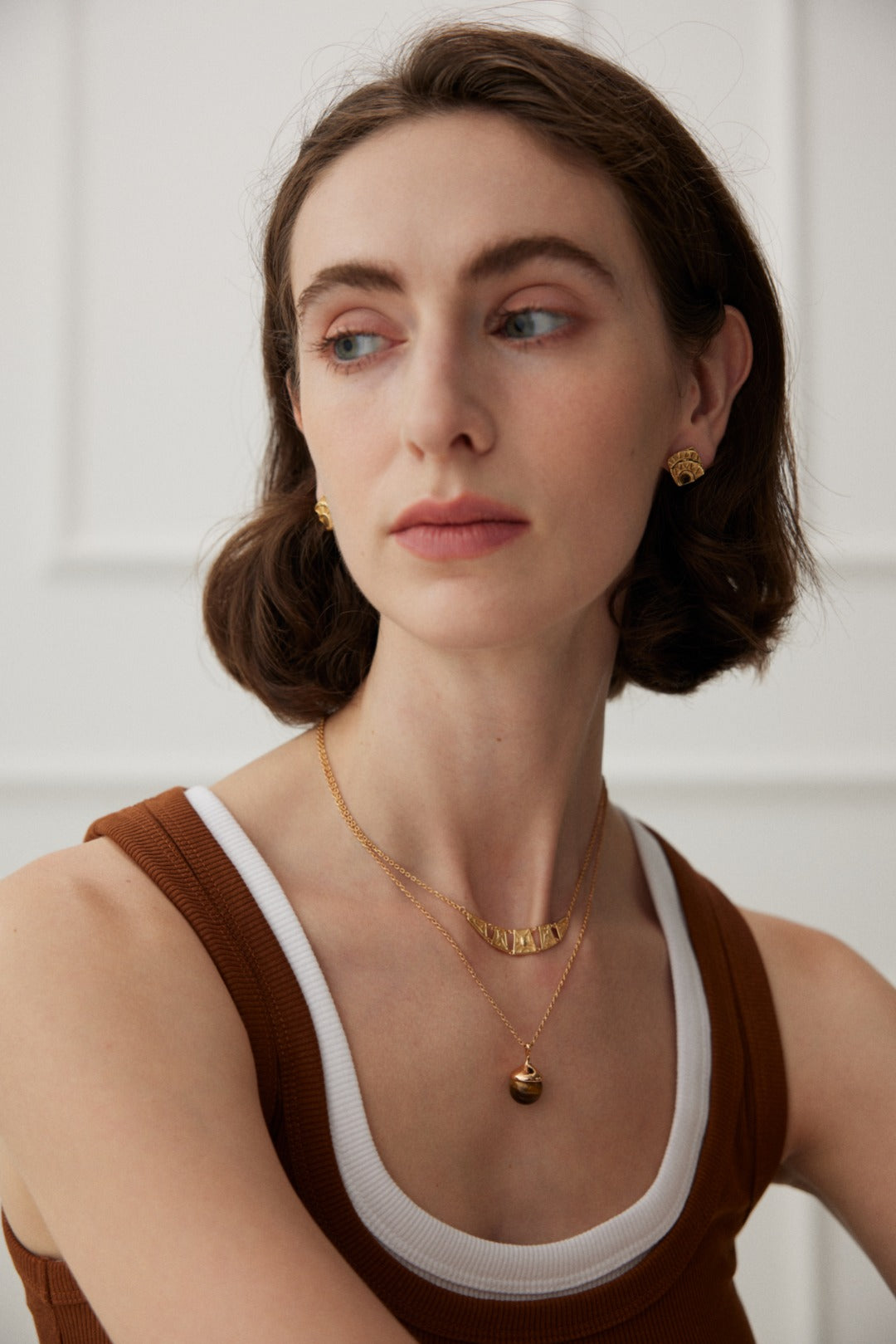 A woman with short brown hair is wearing a brown and white tank top adorned with a subtle geometric pattern. She has on the Vintage Gold Geometric Stud Earrings by Siren Mode and two gold necklaces, one with a pendant and the other featuring small engraved letters. She is looking slightly to her left with a neutral expression.