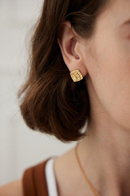 Close-up of a person wearing Siren Mode's Vintage Gold Geometric Stud Earrings with an intricate pattern. Their short, wavy brown hair frames the ear adorned by the jewelry. A glimpse of their neck and a portion of their brown top are visible. The design seamlessly blends modern elegance, reminiscent of stud earrings crafted in 925 silver.