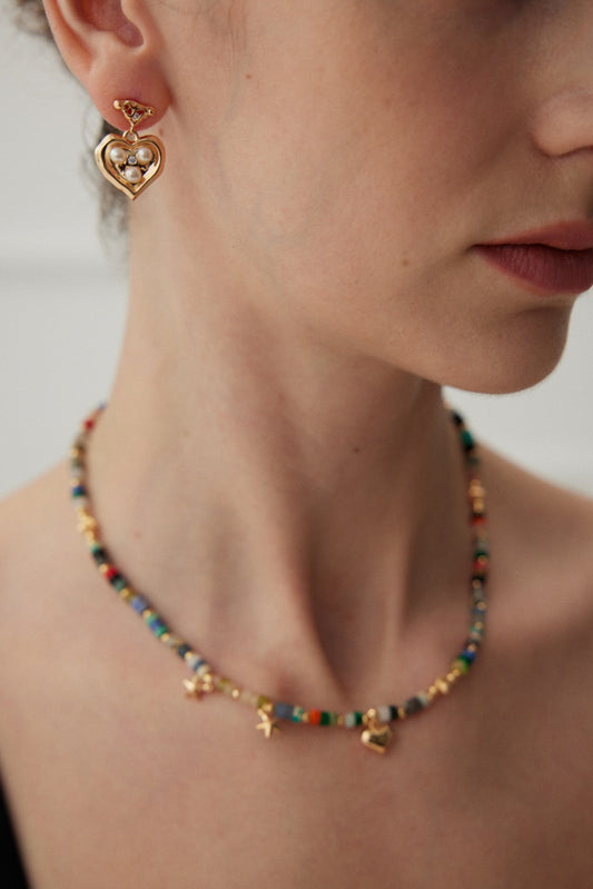 Close-up of a woman's neck adorned with a colorful beaded necklace featuring small charms and natural pearls, complemented by Siren Mode’s Vintage Gold Heart Pearl Earrings. The blurred background beautifully emphasizes the sentimental jewelry. Part of the woman's lips, face, and shoulder are partially visible in the frame.