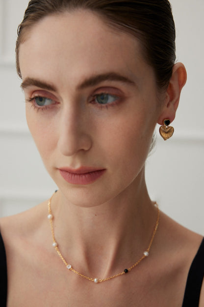 A person with fair skin and slicked-back hair gazes downward. They wear Vintage Gold Heart and Zirconia Earrings from Siren Mode, accented with black, and a delicate gold necklace adorned with small pearls and black beads.