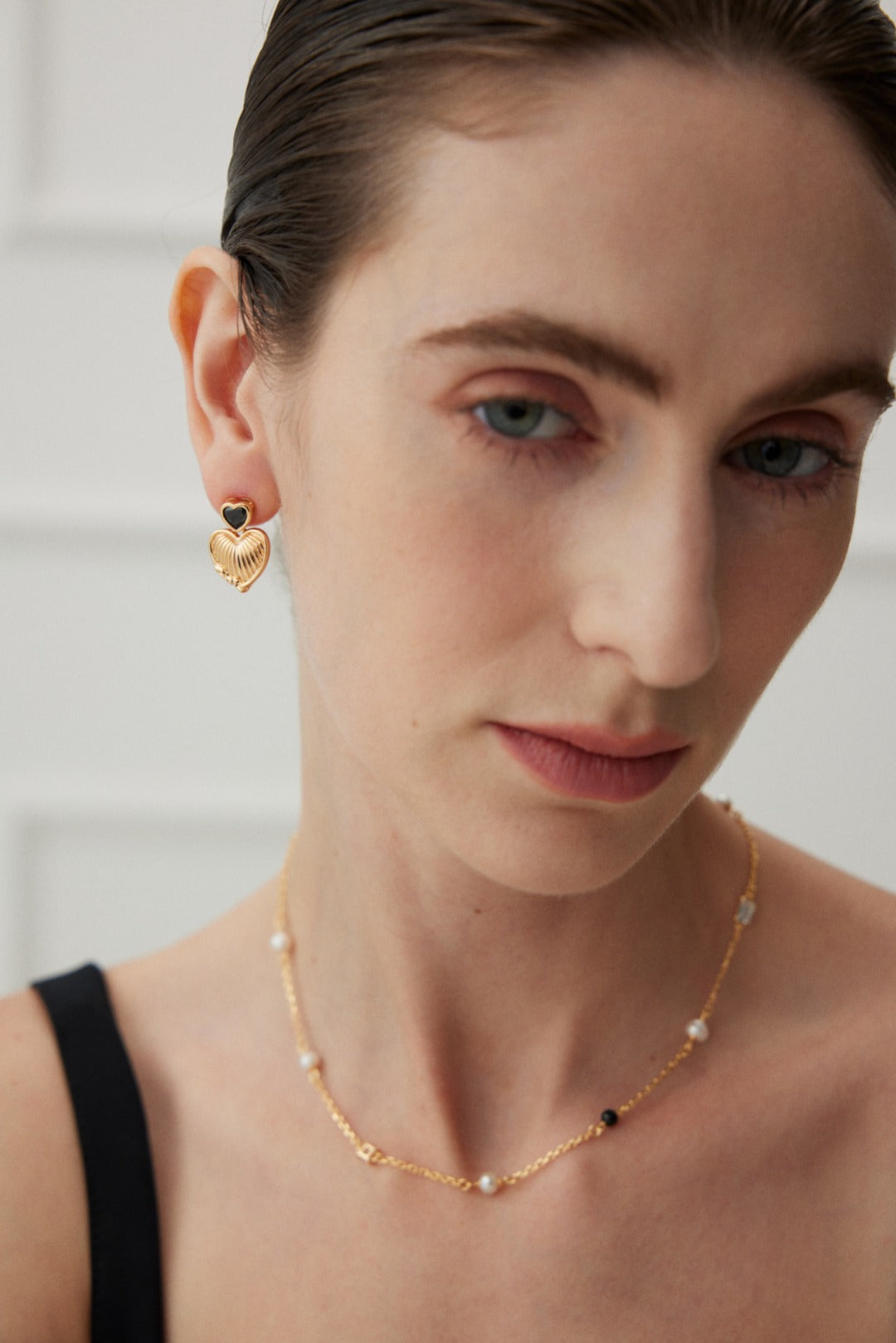 A person with light skin and slicked-back hair is wearing a pair of Vintage Gold Heart and Zirconia Earrings from Siren Mode, along with a delicate gold necklace featuring small pearl-like beads. They have a neutral expression and are dressed in a black top, against a softly blurred background.