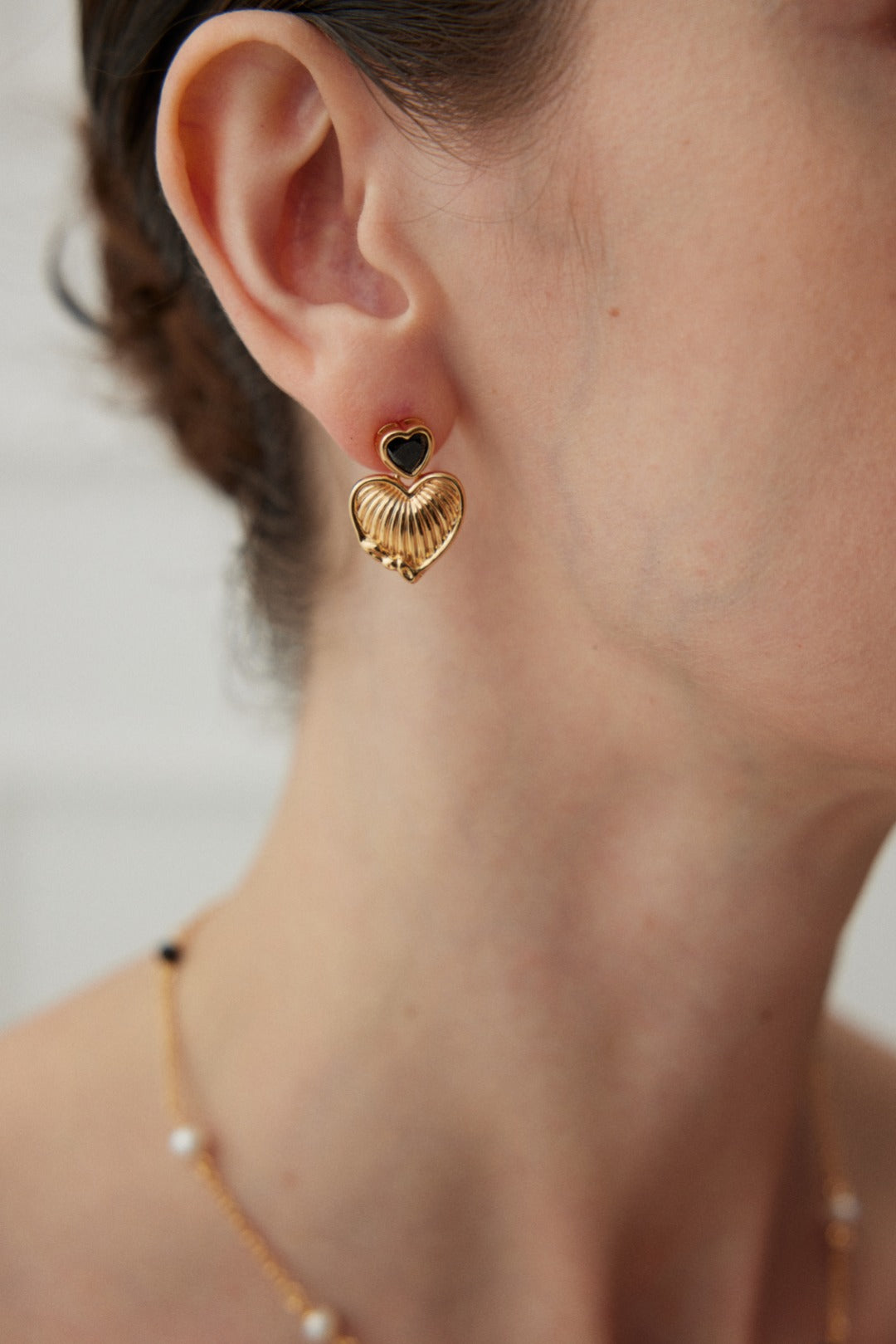 A close-up image of a person's ear adorned with Siren Mode's Vintage Gold Heart and Zirconia Earrings, featuring a small black heart at the top and enhanced by a vintage gold finish. The person's hair is pulled back, and they are wearing a delicate necklace with pearls.