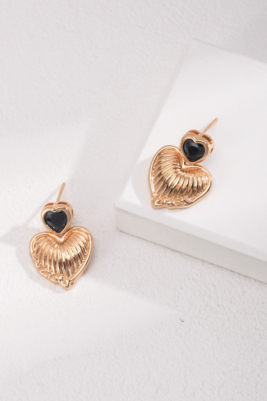 A pair of Siren Mode Vintage Gold Heart and Zirconia Earrings are shown, featuring a black heart at the top connected to a larger, gold textured heart below. Adorned with a vintage gold finish, these earrings are placed on a white surface with a slight shadow cast on the left side.