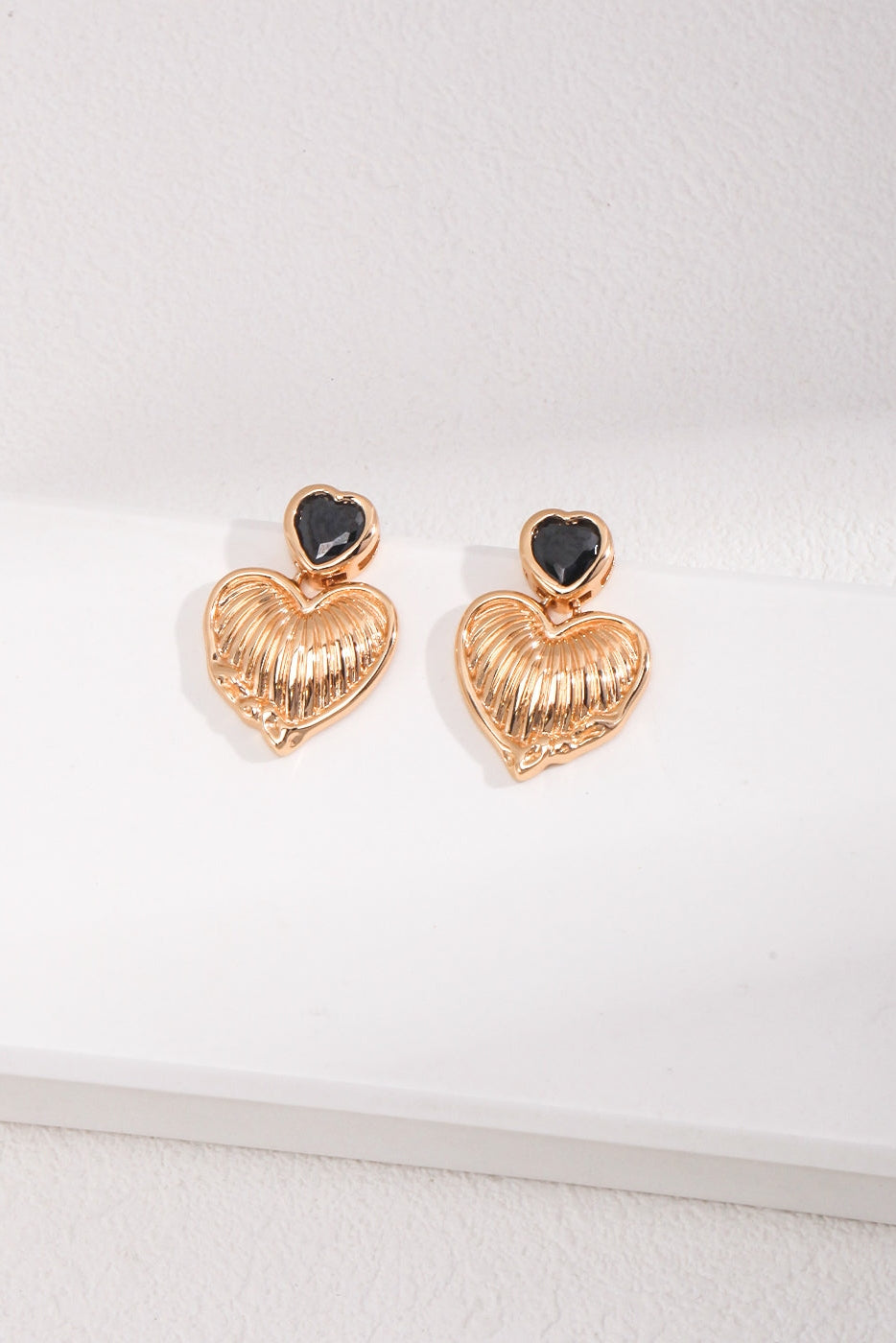 A pair of Siren Mode's Vintage Gold Heart and Zirconia Earrings, with black heart-shaped tops each adorned with a shimmering zirconia stone, are displayed on a white surface. The gold hearts feature a textured, ridged pattern.