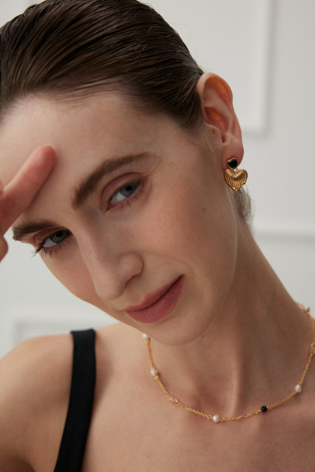 A person with fair skin and brown hair is shown from the shoulders up against a light background. They are wearing a black top, Siren Mode's Vintage Gold Heart and Zirconia Earrings, and a delicate gold necklace. They are gently touching their forehead with two fingers, looking into the camera.