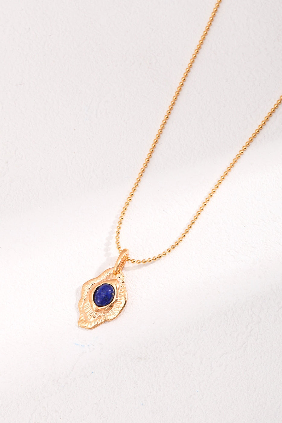 The Vintage Gold Lapis Lazuli Leaf Pendant Necklace by Siren Mode is a delicate gold necklace with a fine chain and an intricate vintage pendant shaped like a leaf or feather. The pendant features an oval-shaped, deep blue lapis lazuli stone set in the center, adding a touch of elegance and color to this artistic jewelry piece.