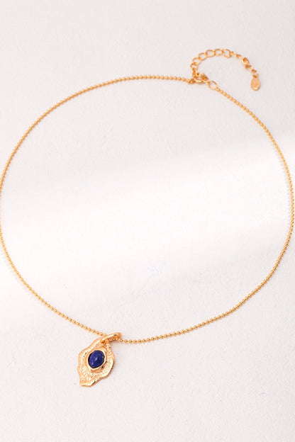 The Siren Mode Vintage Gold Lapis Lazuli Leaf Pendant Necklace is a delicate piece featuring a thin gold chain and a small, ornate leaf-shaped pendant adorned with intricate designs. At the center of the pendant lies a deep blue lapis lazuli stone. This artistic jewelry piece is showcased on a pale surface with subtle lighting.