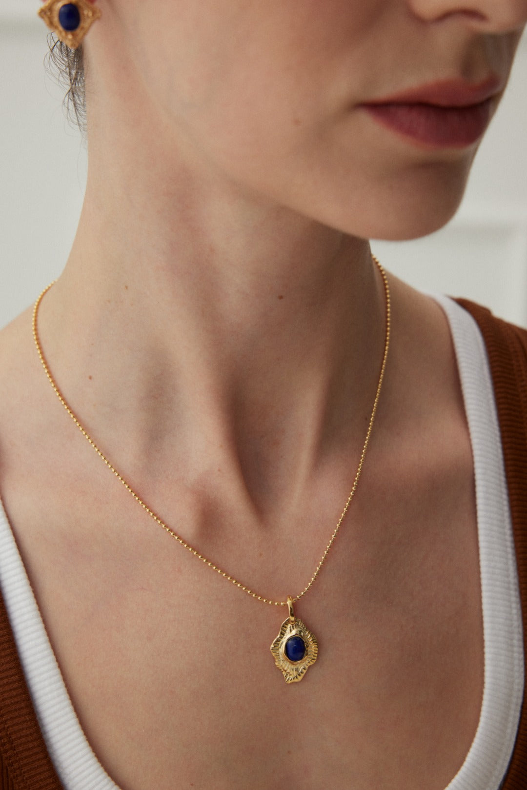 Close-up of a person wearing a Siren Mode Vintage Gold Lapis Lazuli Leaf Pendant Necklace, adorned with blue gemstone earrings. The artistic jewelry complements their white top and brown overlayer, while only the lower part of their face and neck are visible.