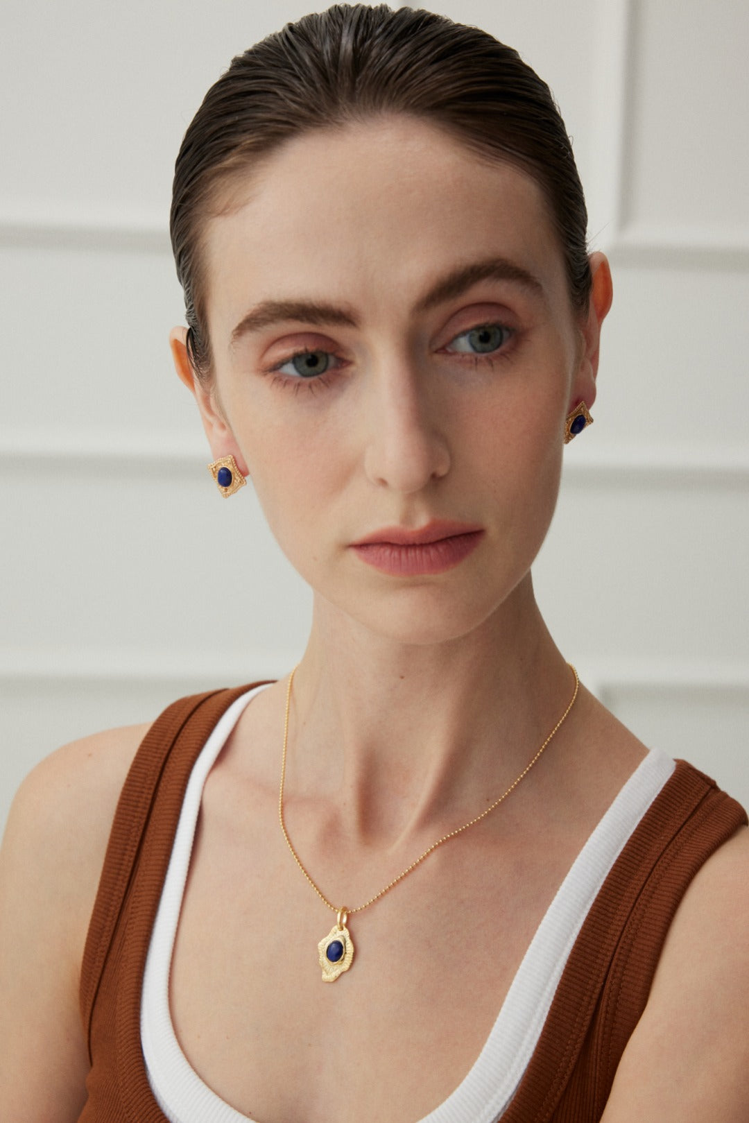 A person with fair skin and slicked-back dark hair is wearing a brown sleeveless top over a white underlayer. They accessorize with artistic jewelry, including the Vintage Gold Lapis Lazuli Leaf Pendant Necklace by Siren Mode along with matching gold earrings adorned with blue stones. The background is plain off-white wall paneling.