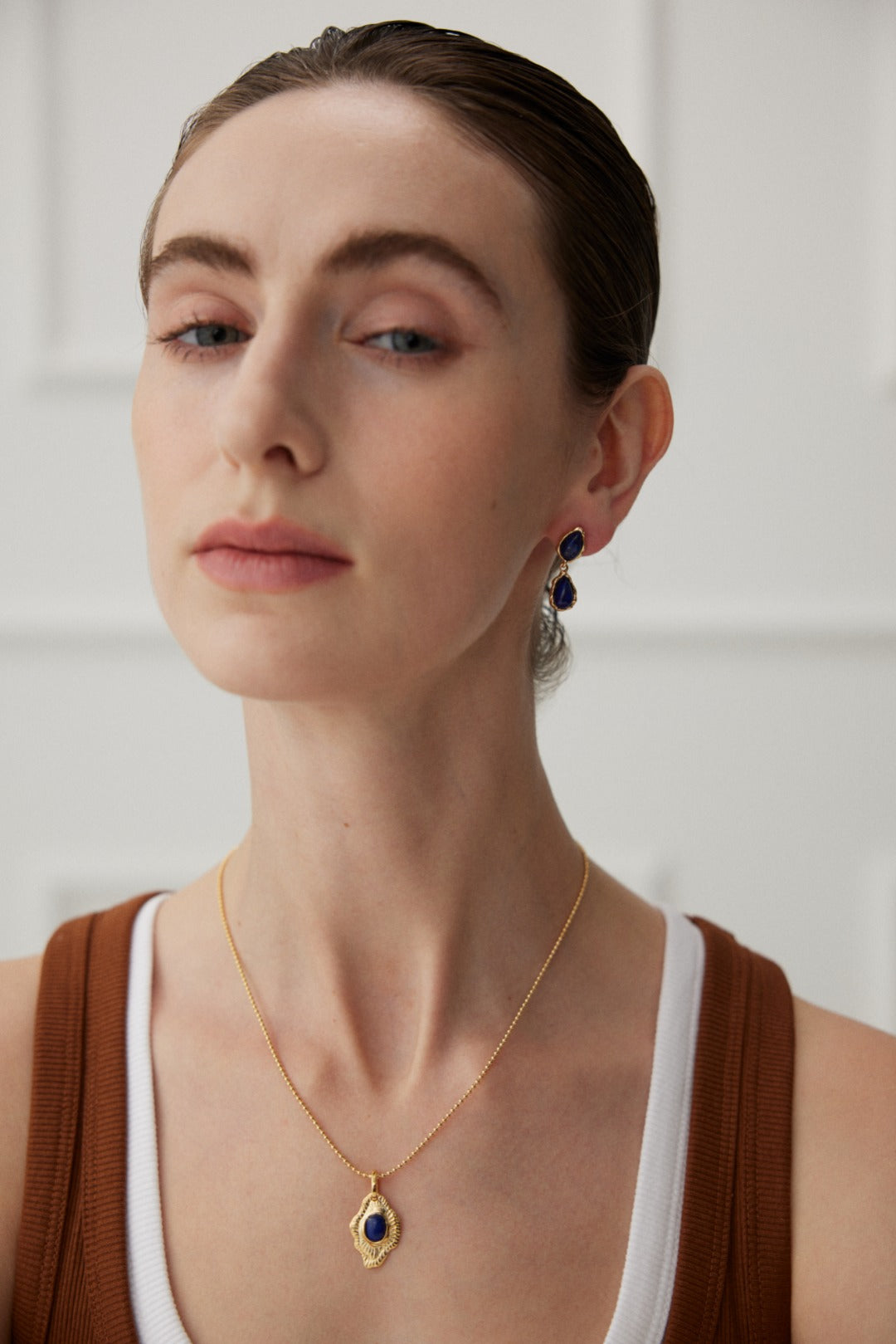 A person with a neutral expression is wearing artistic jewelry from Siren Mode, including a Vintage Gold Lapis Lazuli Leaf Pendant Necklace and matching earrings, both featuring blue stones. They have light skin and styled hair and are dressed in a brown and white outfit. The background is softly blurred.