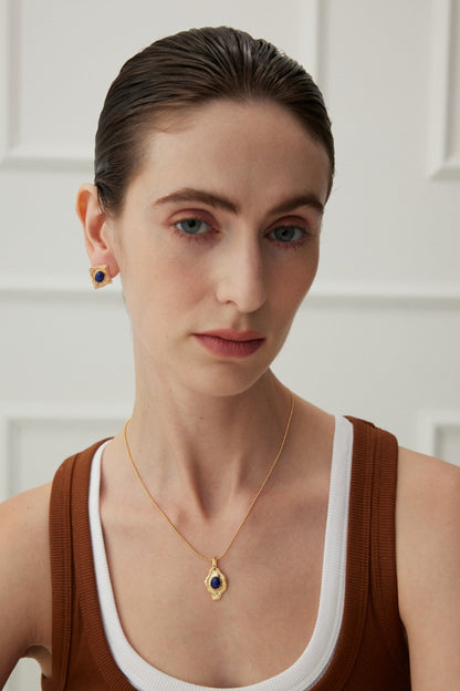 A person with light skin, slicked-back dark hair, and blue eyes wears a brown tank top over a white one. With a serious expression, they accessorize with matching Siren Mode Vintage Gold Lapis Lazuli Leaf Pendant Necklaces. The background showcases a white paneled wall.