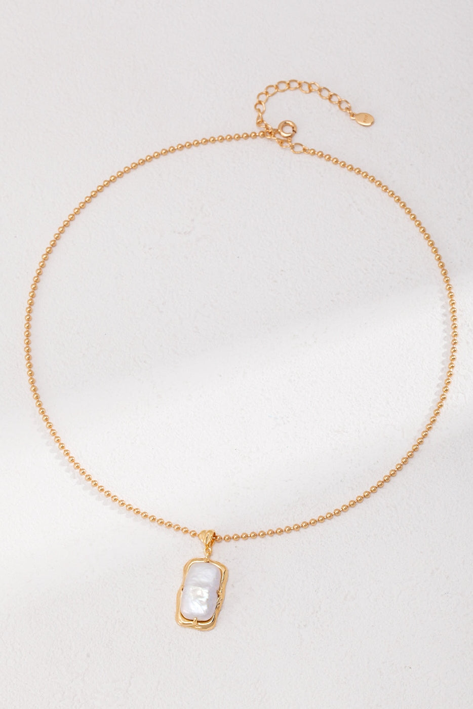 The Vintage Gold Mother of Pearl Pendant Necklace in 925 Silver by Siren Mode is a refined piece featuring a delicate rectangular pendant adorned with a white, textured Mother of Pearl stone. Its adjustable clasp guarantees a perfect fit, allowing the pendant to elegantly hang at the center, framed in gold.