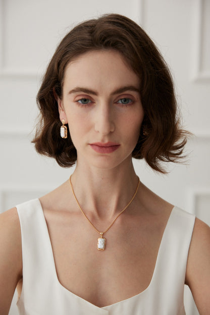 A woman with shoulder-length brown hair wears a white sleeveless top. She has matching rectangular gold earrings and an elegant Vintage Gold Mother of Pearl Pendant Necklace in 925 Silver by Siren Mode. The background is softly lit with white paneled walls.