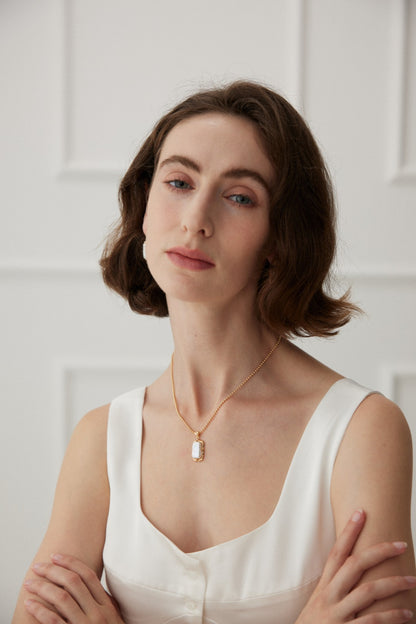 A woman with shoulder-length brown hair is wearing a white sleeveless top and the Vintage Gold Mother of Pearl Pendant Necklace in 925 Silver by Siren Mode. She stands against a simple white paneled background with her arms crossed, looking at the camera with a neutral expression.