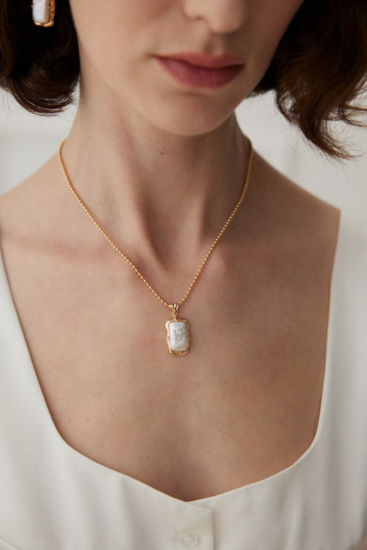 A close-up image of a person wearing the Siren Mode Vintage Gold Mother of Pearl Pendant Necklace in 925 Silver. They have short, wavy dark hair and are dressed in a white, v-neck top with button details. The person's face is partially visible, focusing on their neck and jewelry.