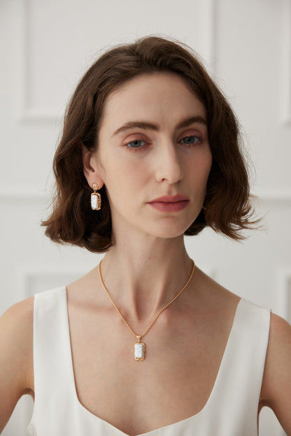 A woman with shoulder-length brown hair is wearing a white sleeveless top. She has on an elegant Siren Mode Vintage Gold Mother of Pearl Pendant Necklace in 925 Silver, paired perfectly with matching rectangular earrings. The background is plain white with subtle panel detailing.