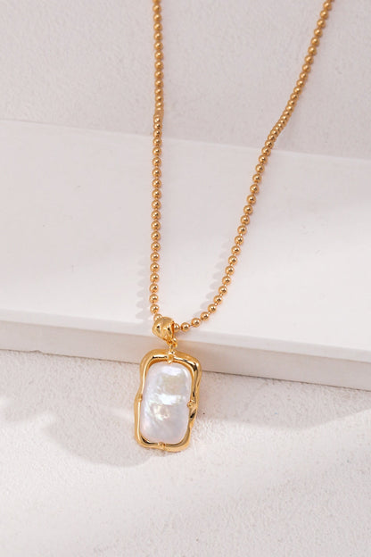 A Vintage Gold Mother of Pearl Pendant Necklace in 925 Silver by Siren Mode is displayed on a white surface. The pendant features a rectangular baroque Mother of Pearl encased in a delicate gold frame. The elegant necklace exudes an elegant and minimalist style.