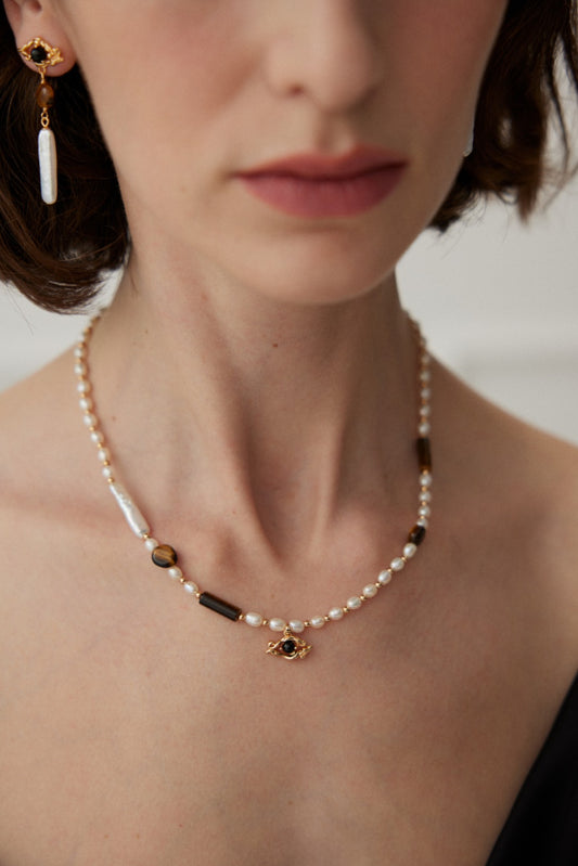 A woman is wearing Siren Mode's vintage gold necklace adorned with natural pearls, Tiger's Eye stones, and black agate accents, accompanied by a matching earring featuring a pearl drop. The image centers on her neck and jewelry, with her face partially visible. She has short, wavy brown hair.