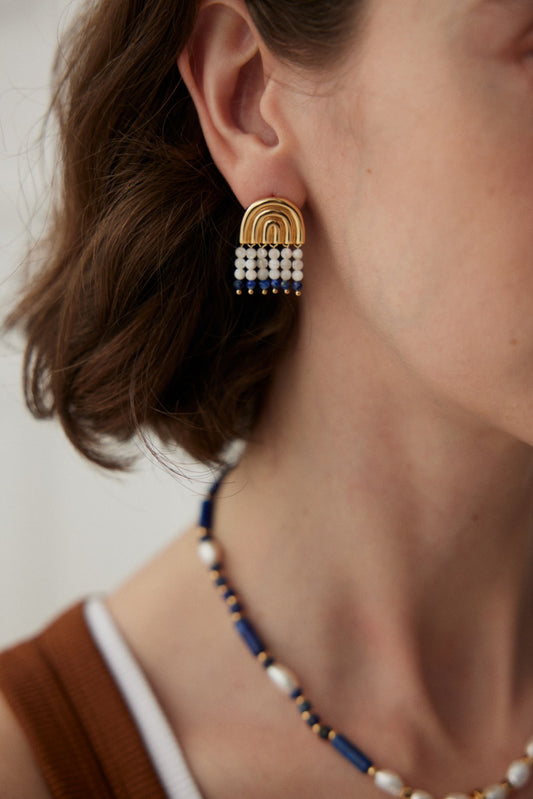 Close-up of a person's ear adorned with Siren Mode's Vintage Gold Rainbow Arch Earrings, featuring delicate Lapis Lazuli and Mother-of-Pearl beads. The person also sports an artful jewelry piece—a necklace made of blue and white beads. The background is out of focus, drawing attention to the exquisite jewelry.