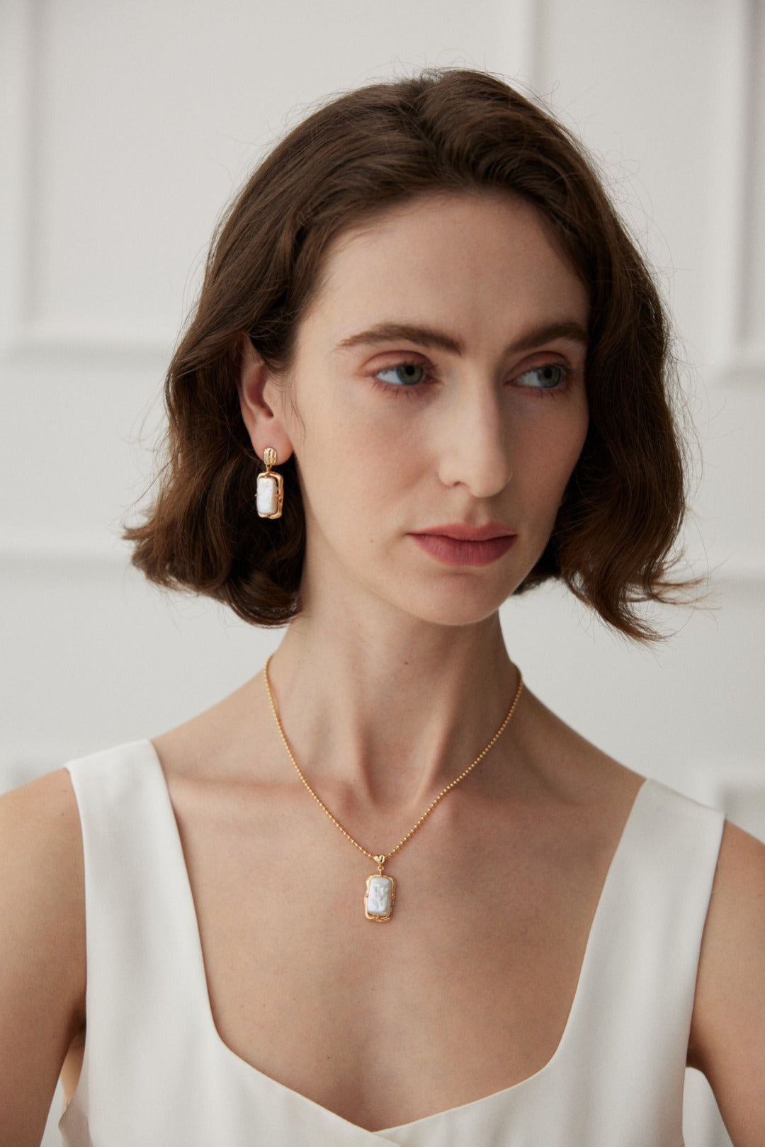 A woman with shoulder-length brown hair is wearing a white sleeveless top. She accessorizes with a vintage gold necklace featuring a rectangular pendant and the "Vintage Gold Rectangular Mother of Pearl Drop Earrings in 925 Silver" by Siren Mode. She looks slightly to the side with a neutral expression against a softly lit white background.