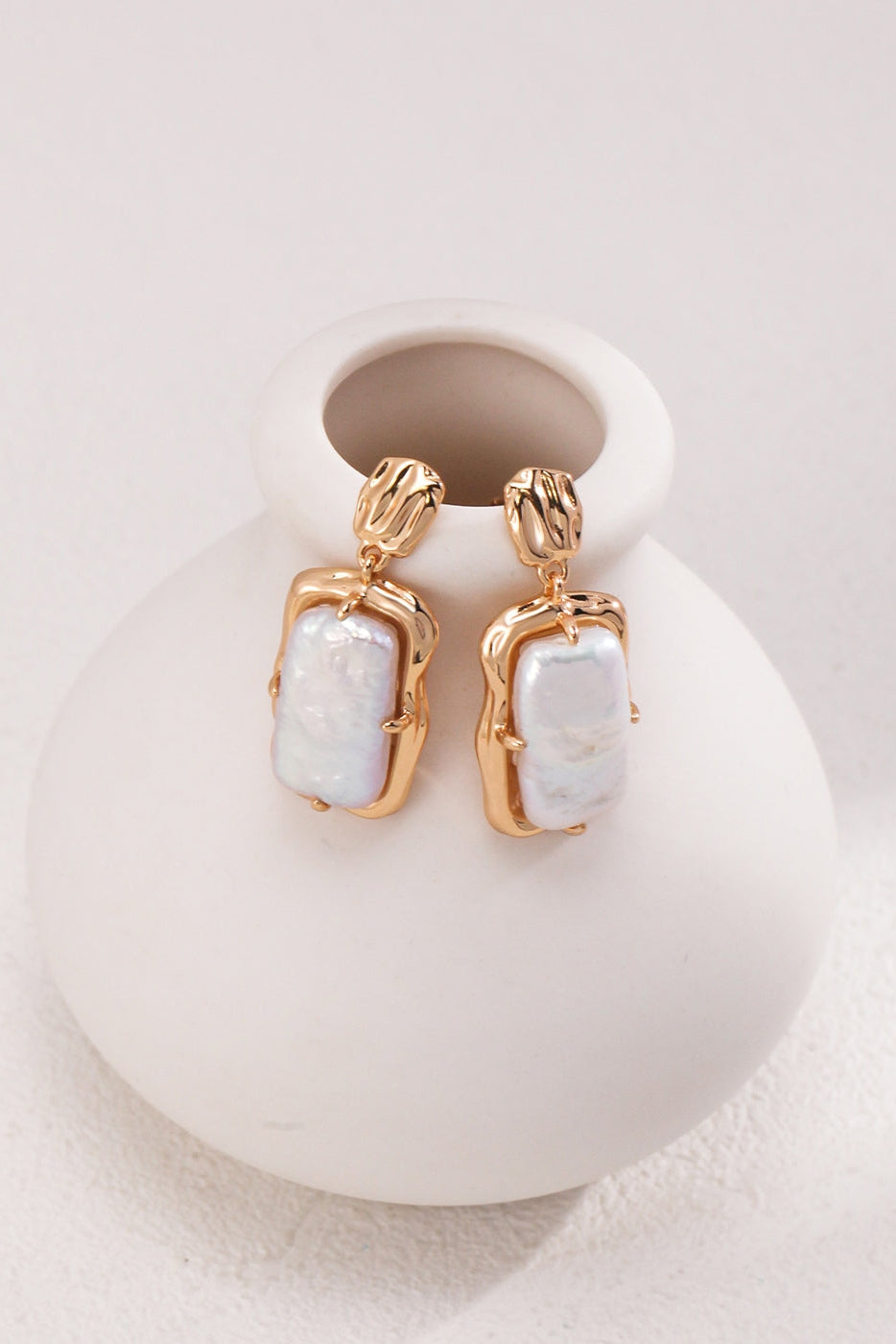 A pair of Siren Mode's Vintage Gold Rectangular Mother of Pearl Drop Earrings in 925 Silver is showcased elegantly hanging from the rim of a smooth, white, round vase. The irregularly shaped Mother of Pearl pieces are set in vintage gold frames with intricate detailing on the earring clasps. The background is a plain, light surface.