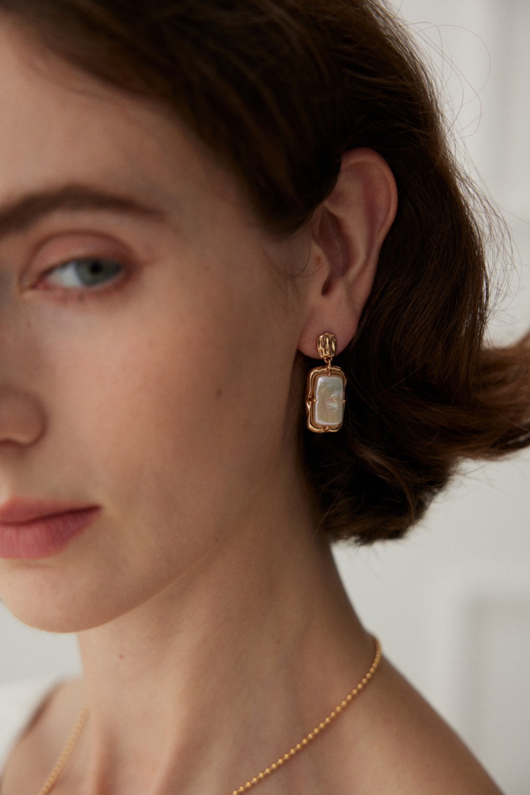 Close-up of a person with fair skin wearing Siren Mode's Vintage Gold Rectangular Mother of Pearl Drop Earrings in 925 Silver and a delicate gold necklace. The earrings showcase a rectangular design with a mother of pearl centerpiece encased in a gold frame. The person's short, brown hair is visible, and their head is slightly turned.