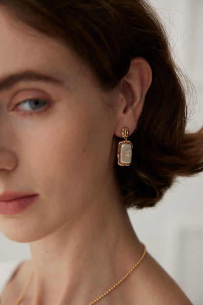 Close-up of a person with fair skin wearing Siren Mode's Vintage Gold Rectangular Mother of Pearl Drop Earrings in 925 Silver and a delicate gold necklace. The earrings showcase a rectangular design with a mother of pearl centerpiece encased in a gold frame. The person's short, brown hair is visible, and their head is slightly turned.