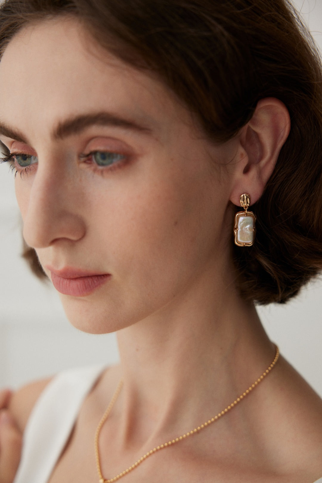 A woman with short brown hair is dressed in a white top and adorned with vintage gold jewelry. She wears a delicate mother of pearl necklace and a pair of Siren Mode's Vintage Gold Rectangular Mother of Pearl Drop Earrings in 925 Silver. The photo highlights her face and the left side of her head against a soft background.