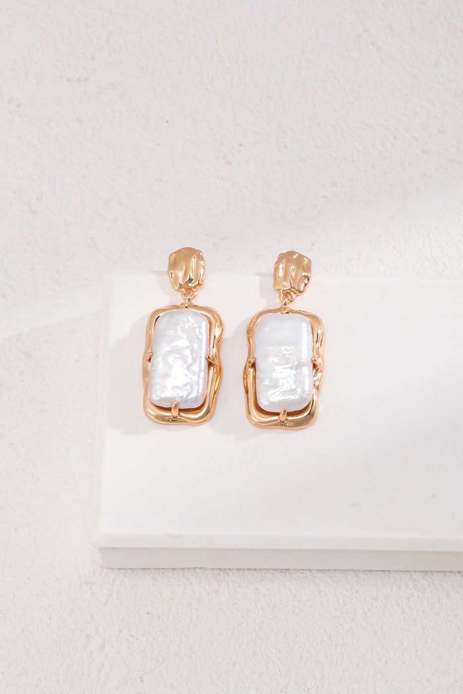 A pair of Siren Mode Vintage Gold Rectangular Mother of Pearl Drop Earrings in 925 Silver are displayed on a light-colored surface. The earrings feature a modern, minimalist design with smooth edges and a polished finish, set against a textured neutral-toned background.