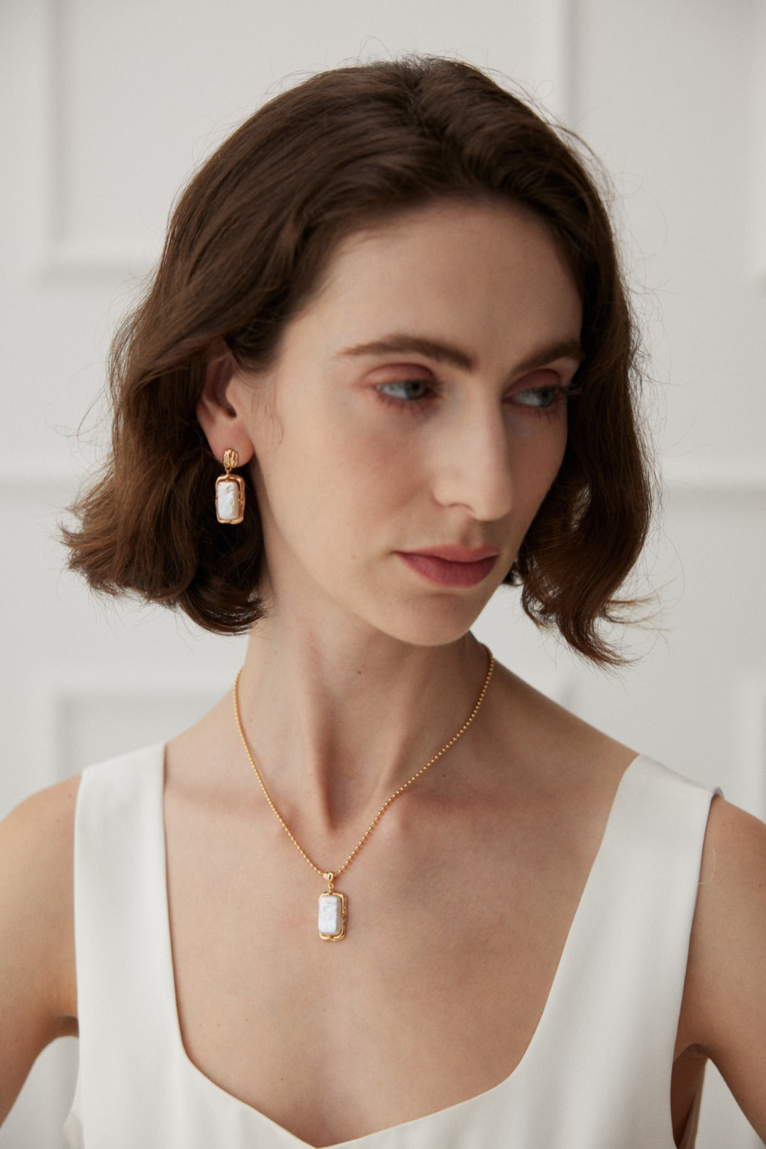 A woman with short brown hair is wearing a white sleeveless top. She complements her look with Siren Mode Vintage Gold Rectangular Mother of Pearl Drop Earrings in 925 Silver and a matching vintage gold necklace, both adorned with rectangular white stones. The background features a white wall with rectangular paneling.