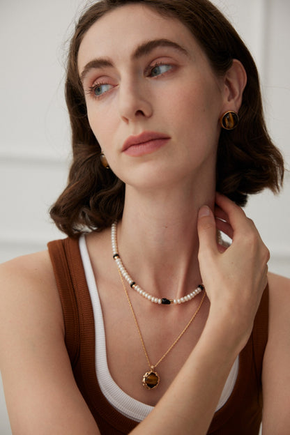 A woman with short brown hair is looking to the side. She is wearing a brown tank top with a white layer underneath. She has on two necklaces: one crafted from semi-precious stones and pearls, and another featuring a circular gold pendant; she also wears matching earrings named "Vintage Gold Tiger's Eye Stud Earrings" by Siren Mode.