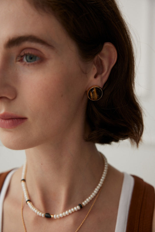 A close-up shows a woman with short brown hair wearing the Vintage Gold Tiger's Eye Stud Earrings by Siren Mode and a layered necklace featuring pearls, black beads, and semi-precious stones. She dons a brown and white top. Her blue eyes and light skin are set against a neutral background.