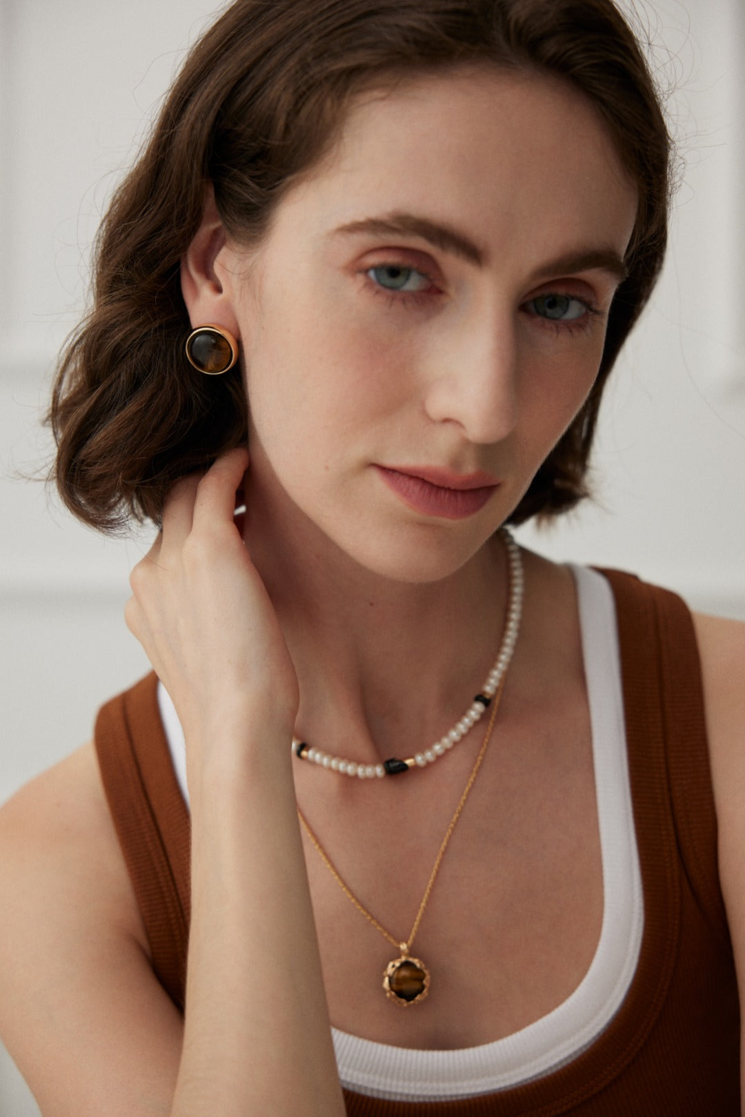 A woman with short brown hair wears a brown sleeveless top over a white shirt. She accessorizes with Siren Mode's Vintage Gold Tiger's Eye Stud Earrings, a pearl necklace, and a layered gold necklace featuring Tiger's eye gemstones. Her hand touches her neck as she looks softly into the camera.