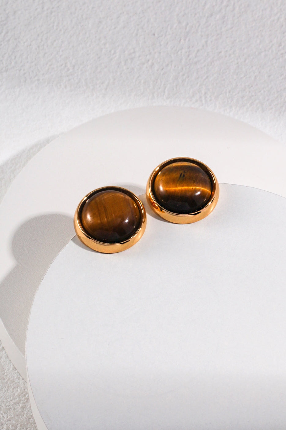 A pair of Vintage Gold Tiger's Eye Stud Earrings by Siren Mode rests on a circular white surface. The semi-precious tiger's eye gemstones in the gold-framed earrings display their characteristic brown and golden streaks.