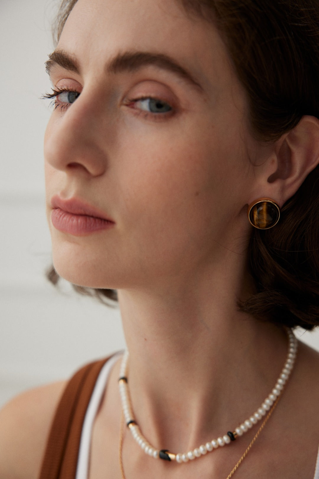 A close-up shot features a light-skinned individual with brown hair, gazing slightly to the side. They adorn a pearl necklace interspersed with small black beads and are wearing Siren Mode's Vintage Gold Tiger's Eye Stud Earrings. The softly blurred, light-colored background adds an elegant touch to the scene.