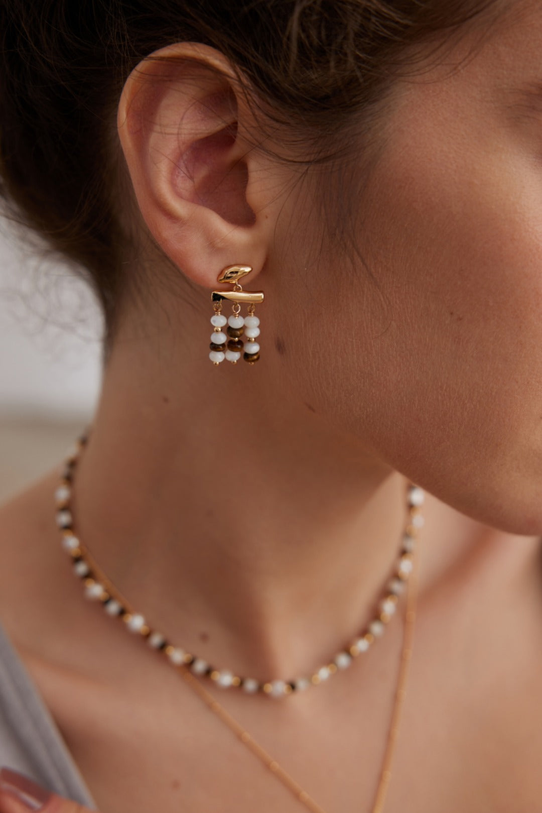 Close-up of a person's ear adorned with Siren Mode's Vintage Gold Tiger's Eye and Mother of Pearl Drop Earrings in 925 Silver, featuring three dangling beaded strands in white and black. They are also wearing a matching beaded necklace with similarly colored beads. The person has light brown hair tied back.