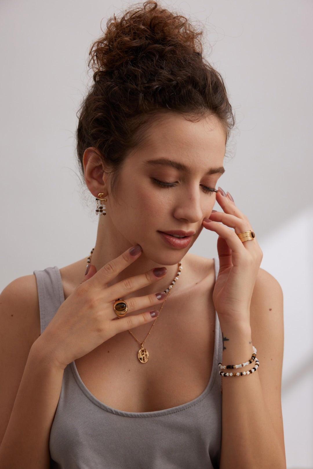 A woman with curly hair in a bun touches her face and neck, wearing a sleeveless gray top. She wears various pieces of jewelry, including Mother of Pearl rings, a Vintage Gold bracelet, and a necklace. Her eyes are gently closed, and she exudes a serene and contemplative expression. Adorning her ears are the elegant Siren Mode Vintage Gold Tiger's Eye and Mother of Pearl Drop Earrings in 925 Silver.