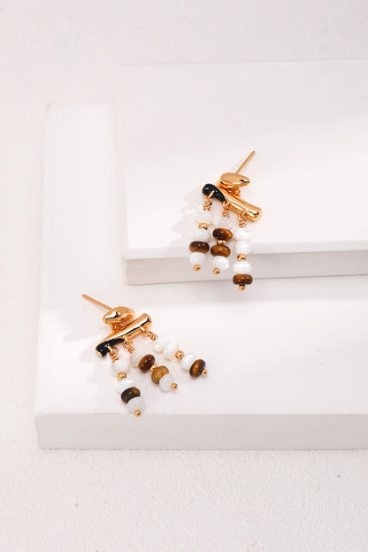 Vintage Gold Tiger's Eye and Mother of Pearl Drop Earrings in 925 Silver by Siren Mode, featuring dangling multicolored beads in shades of brown, white, and black, are displayed on minimalist white blocks, creating a stylish and elegant presentation.