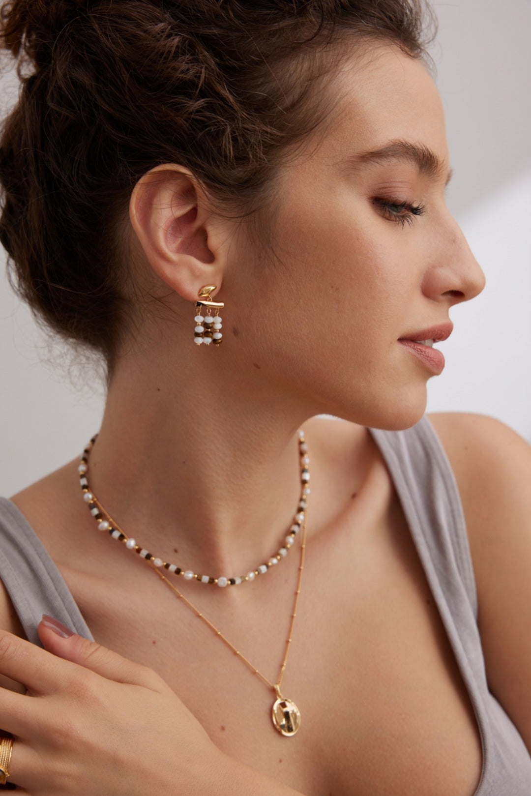 A woman is photographed in a profile view wearing a light gray tank top. She has on Siren Mode's Vintage Gold Tiger's Eye and Mother of Pearl Drop Earrings in 925 Silver, a double-layer pearl and gold bead necklace, and another gold necklace with a circular pendant. Her hair is styled in a messy bun.