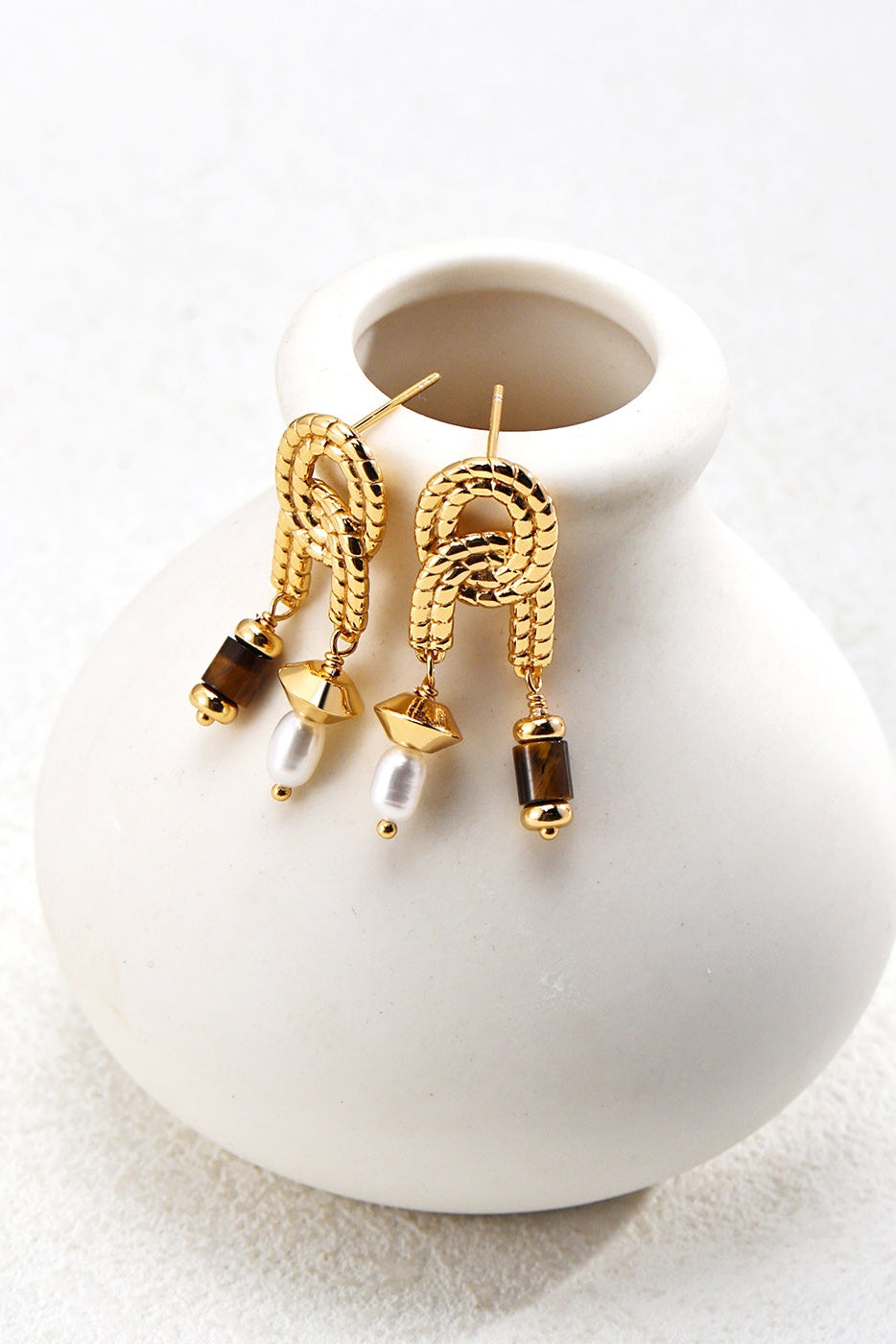 A pair of Vintage Gold Tiger's Eye and Pearl Drop Earrings by Siren Mode gracing the rim of a small white vase. These elegant earrings showcase an arched design, each adorned with a black bead and a natural pearl, imparting a sophisticated and stylish look.